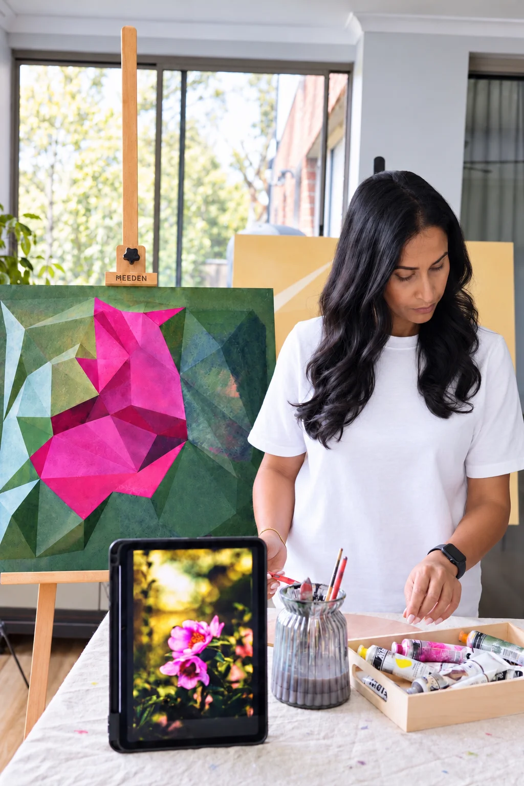 Magenta and green artwork on a studio easel beside a person, with a flower reference displayed on a tablet